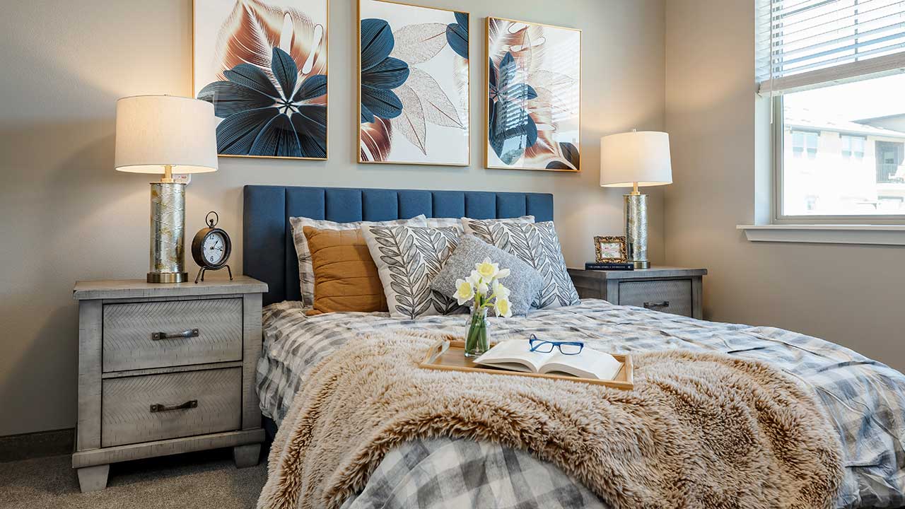 A cozy bedroom with a plaid bedspread, decorative pillows, and a fur throw. A tray with an open book, glasses, and flowers sits on the bed. Two bedside tables with lamps flank the bed, and three floral artworks hang on the wall above the headboard.