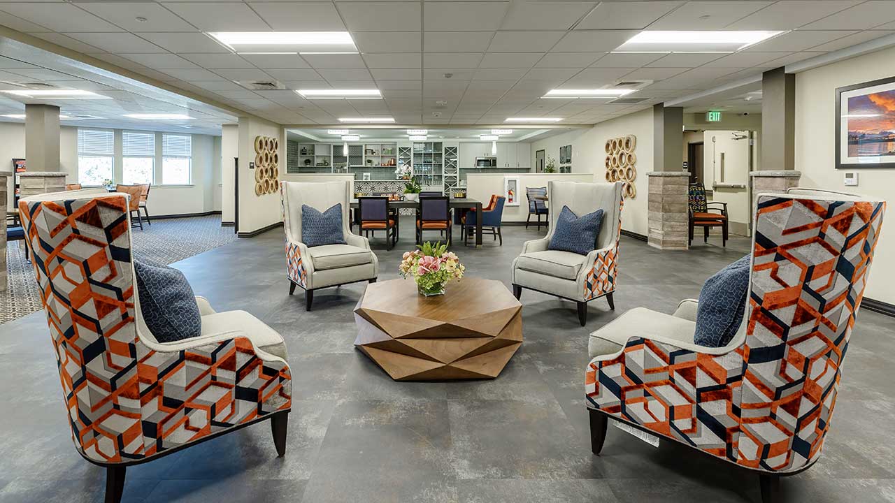 A modern, spacious lounge featuring geometric-patterned armchairs around a wooden coffee table with a floral centerpiece. In the background, a dining area and a corridor are visible. Natural light streams in through large windows.