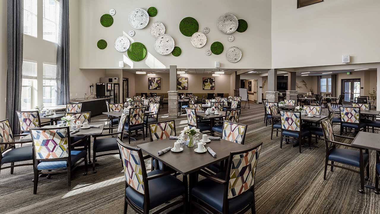 A spacious dining area with several tables and chairs, each set with white tableware. The walls are adorned with abstract decorations and green plant accents. Large windows allow natural light to fill the room, creating a bright, inviting atmosphere.