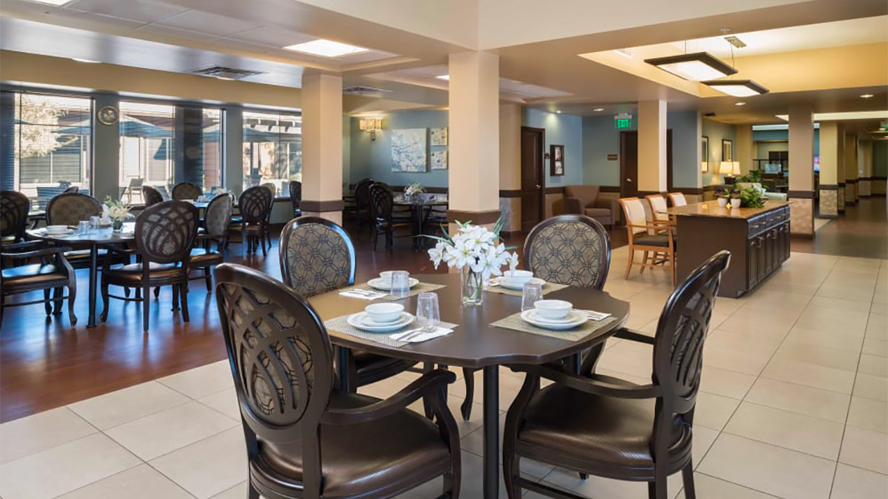 A spacious dining area with elegant wooden tables and chairs. Centerpieces of white flowers are on each table. The room has large windows, soft lighting, and framed artwork on the walls, creating a warm and inviting atmosphere.