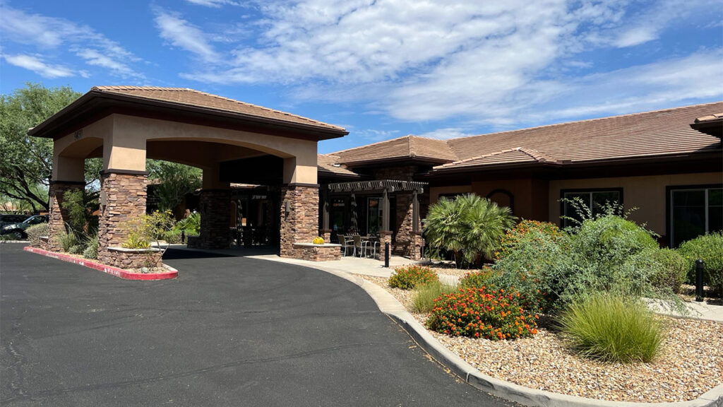 A building with a covered entrance supported by stone columns, surrounded by landscaped gardens with shrubs and colorful flowers. The sky is partly cloudy, and there is a paved driveway leading to the entrance.