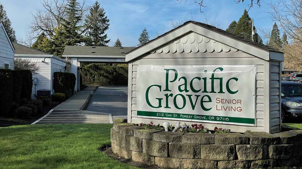 Sign reading "Pacific Grove Senior Living" in front of a well-maintained residential building with a driveway and manicured landscaping. The sign includes an address: 2112 Oak St., Forest Grove, OR 97116. Trees and shrubs are in the background.