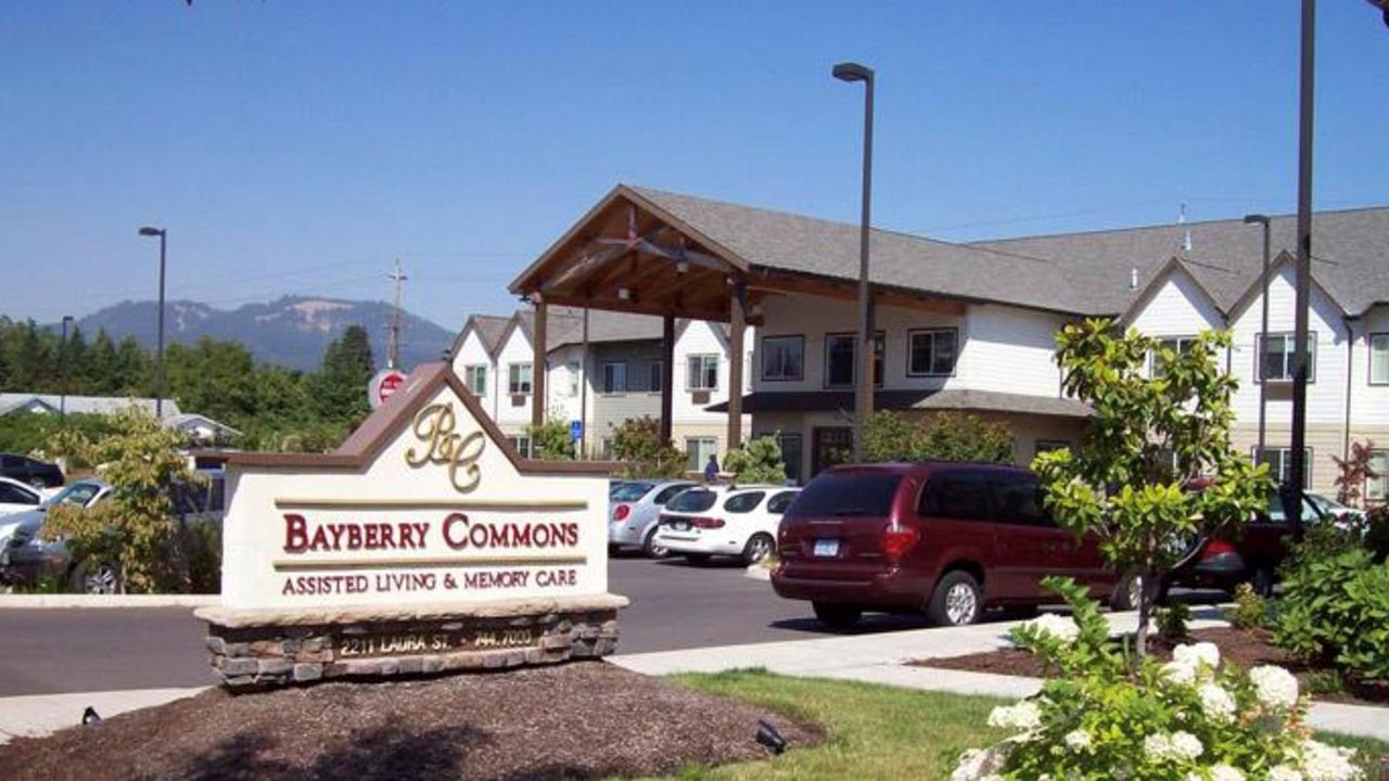 Image of Bayberry Commons Assisted Living & Memory Care facility. The foreground shows a sign with the name, surrounded by landscaping. In the background, there is a two-story building and a parking lot filled with cars.