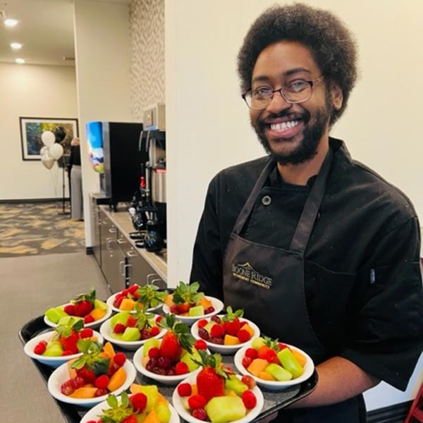 A smiling person in a black chef uniform holds a tray with bowls of fresh fruit, including strawberries, melons, and grapes. Balloons and a coffee station are visible in the background.