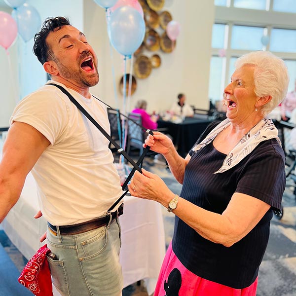 A man playfully grimaces as an elderly woman tugs on his suspenders. Both appear amused and cheerful, surrounded by pastel balloons and natural light streaming through large windows in a festive setting.