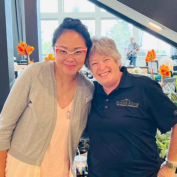 Two women smiling and standing close together. One wears a light pink dress with a gray cardigan and glasses, while the other wears a black polo shirt with the words "Boone Ridge Retirement Community" embroidered. Bright orange flowers are in the background.