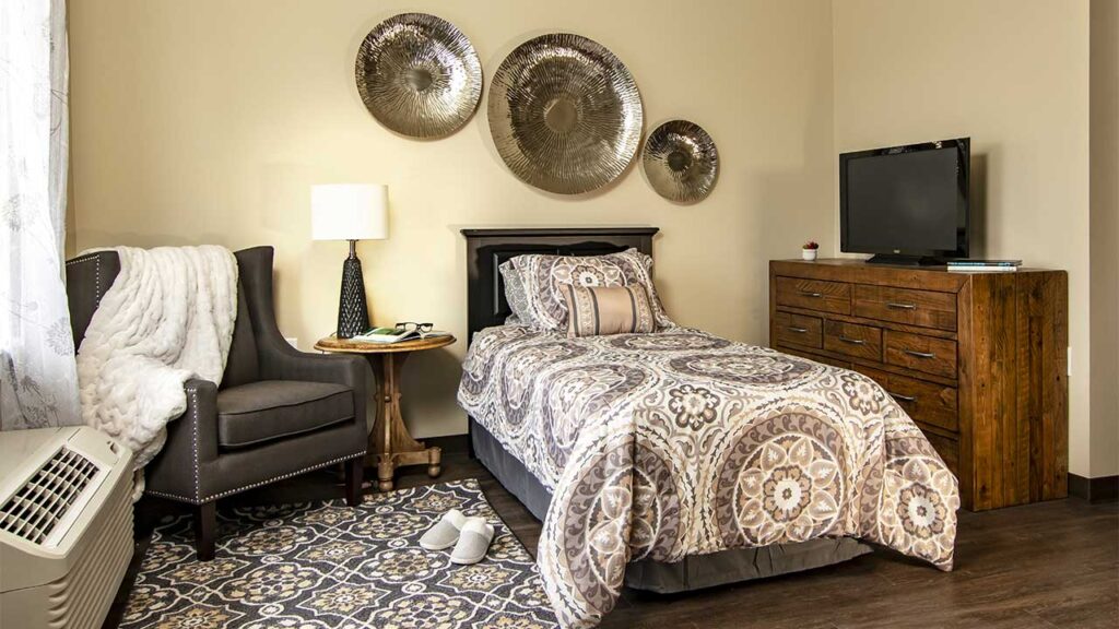 A cozy bedroom with a single bed featuring patterned bedding, a wooden dresser with a TV, a gray armchair, and a lamp on a side table. Three decorative wall plates hang above the bed. A pair of white slippers rests on a patterned rug beside the bed.