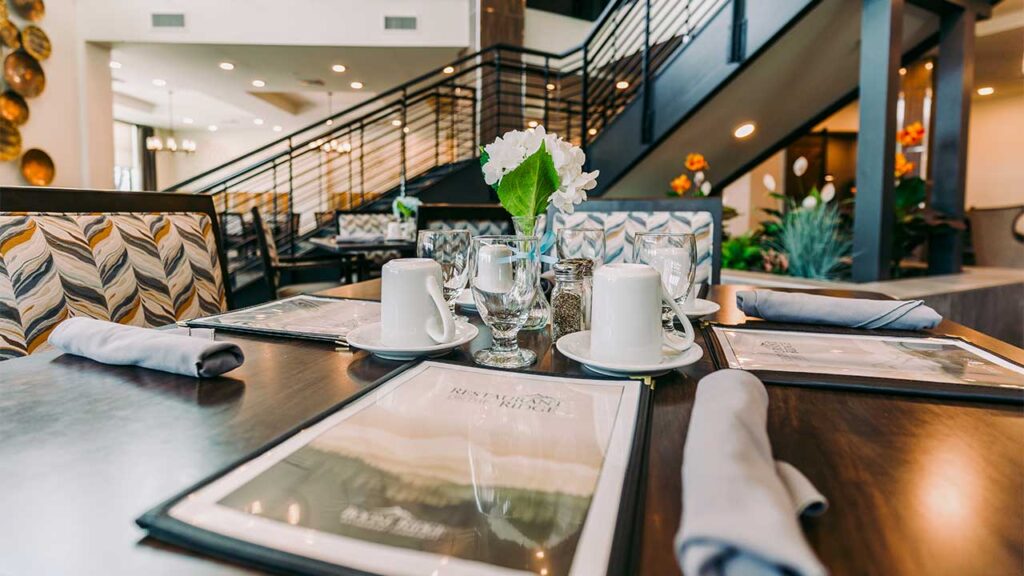 A modern restaurant interior with a neatly set dining table in the foreground. The table includes white mugs, glasses, napkins, and a small flower arrangement. A staircase and decorative plants are visible in the background.