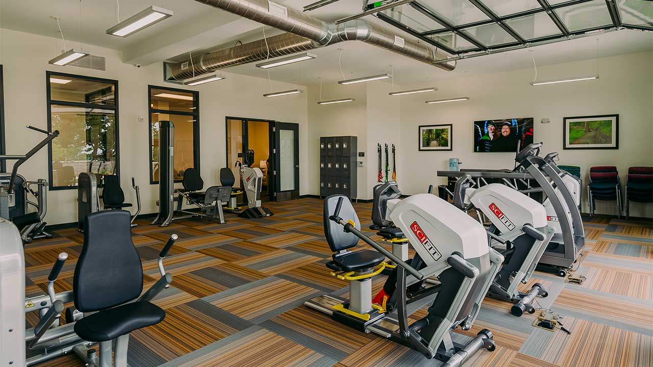 A modern gym with various exercise machines, including stationary bikes and elliptical trainers. The room has large windows, a patterned floor, and a TV mounted on the wall. A person in a white shirt is standing near the entrance.