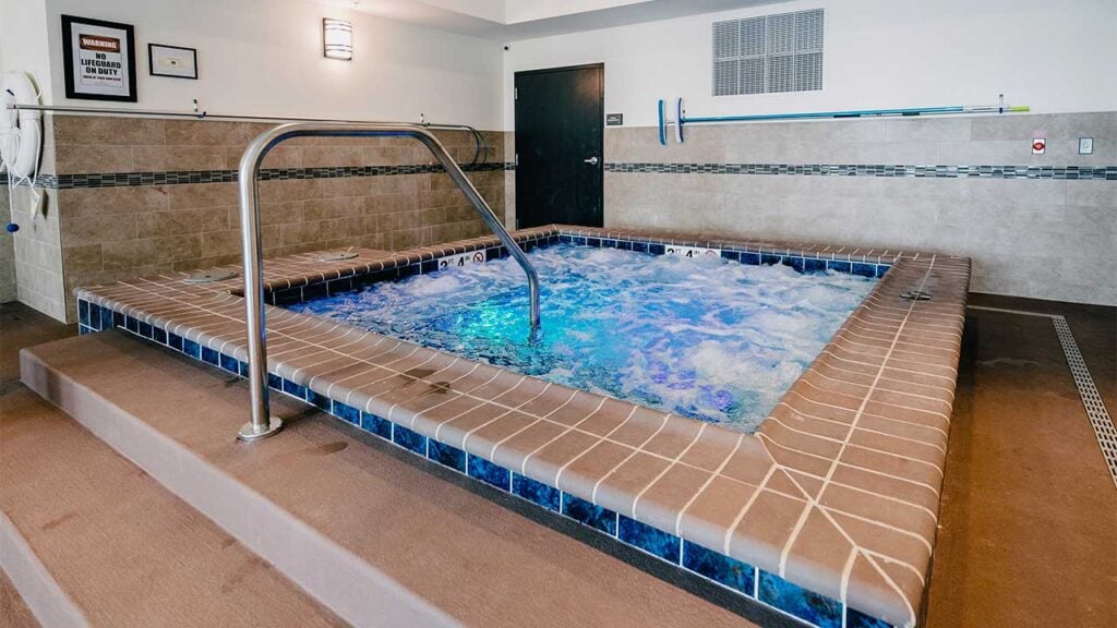 Indoor hot tub with bubbling water surrounded by brown tiles. Walls are tiled with beige and blue accents. A handrail leads into the water, and framed decor hangs on the walls.