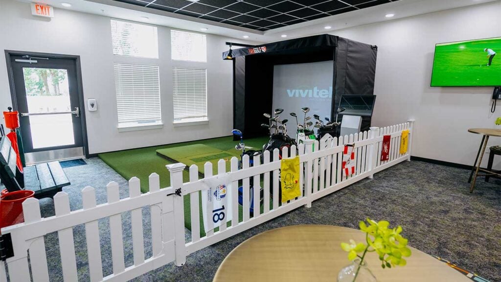 A modern indoor golf setup featuring a golf simulator and various golf clubs. The area is enclosed by a white picket fence, with a video screen displaying golf scenery and a TV mounted on the wall. Bright room with windows and a table with a plant.