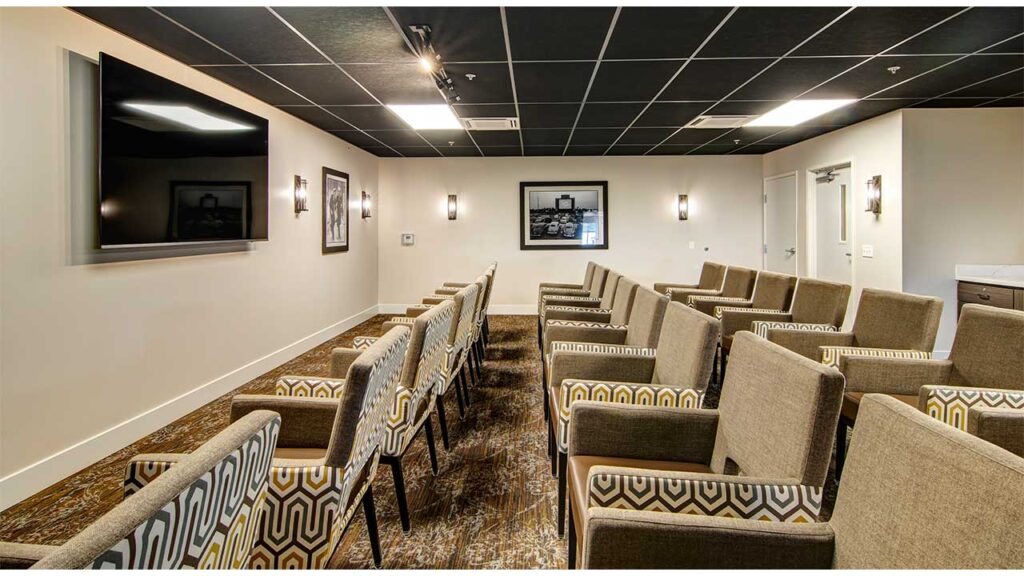 A modern conference room with patterned chairs arranged in rows facing a large wall-mounted TV. The room has framed pictures on the walls, carpeted flooring, and soft lighting from wall sconces. The ceiling is black with recessed lights.