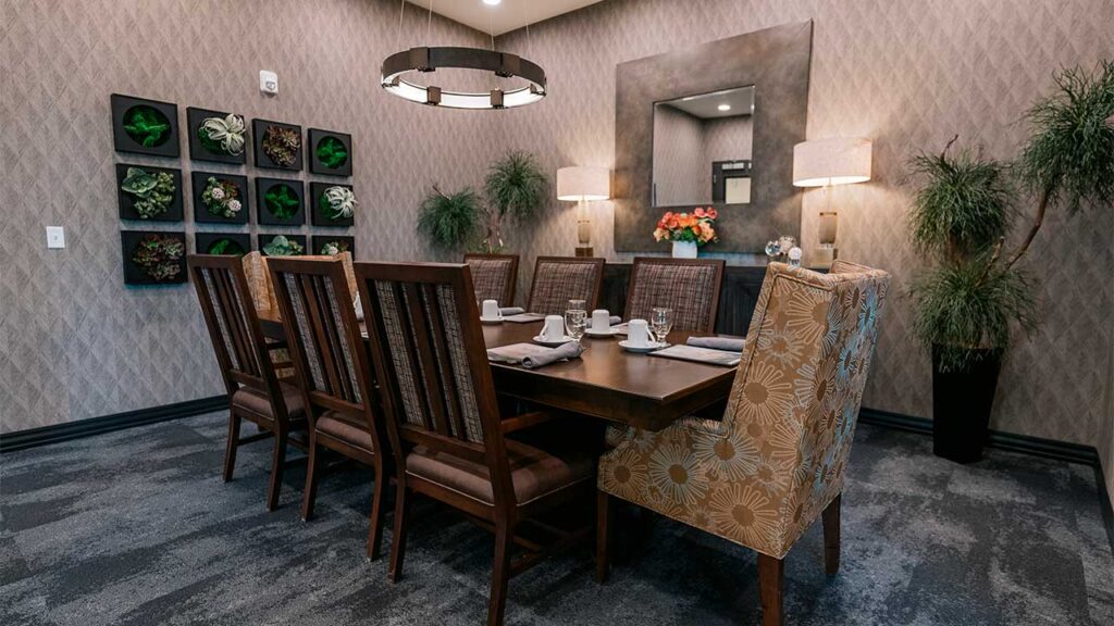 A dining room with a long wooden table surrounded by chairs. The room features a patterned wallpaper, a large mirror, wall art with green plant designs, and warm lighting from an overhead fixture and lamps. There are plates and cups on the table.