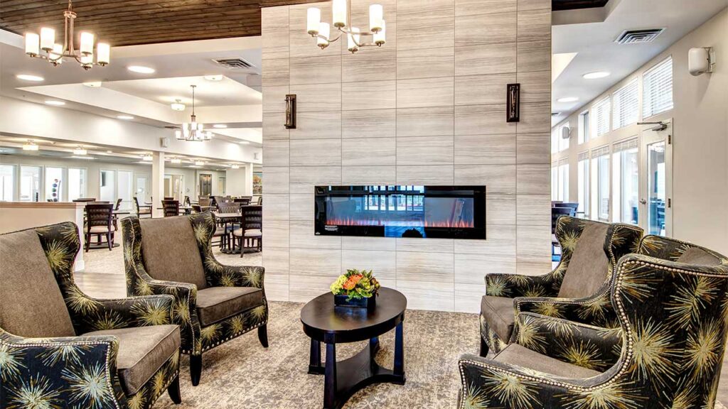 A cozy lounge area with four upholstered armchairs surrounding a small round table with a flower arrangement. A modern electric fireplace is set in a large tile wall. Chandeliers hang from the ceiling, and the room is bright with natural light.