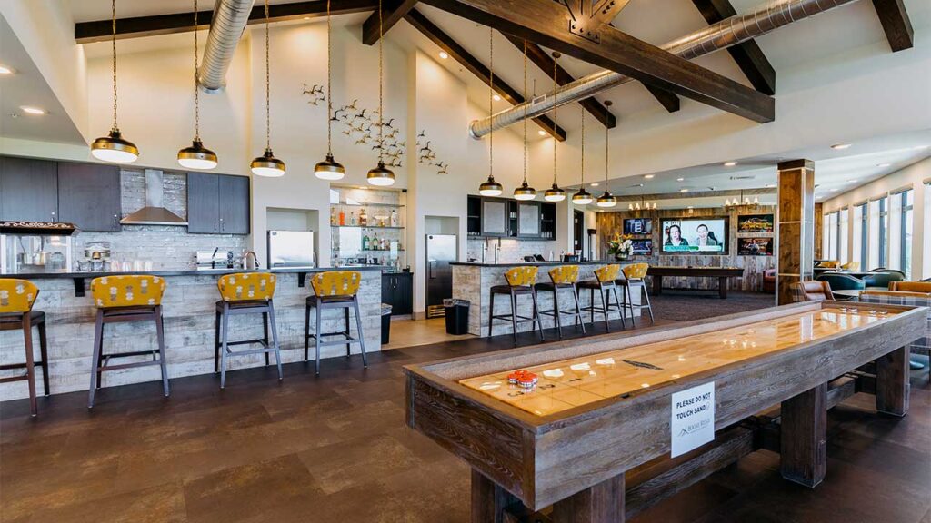 A spacious bar area with wooden accents features a long counter with yellow cushioned bar stools, overhead pendant lights, a shuffleboard table, and multiple TV screens on the wall. Ceiling beams add to the rustic decor.