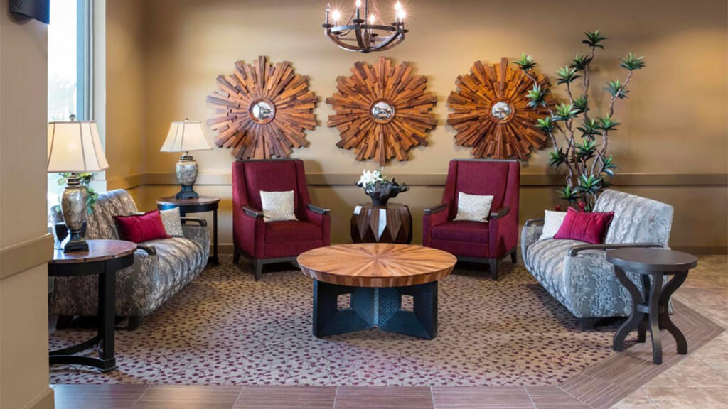A cozy living room with two red armchairs and two patterned sofas arranged around a wooden coffee table. Three decorative wooden wall hangings and a chandelier add warmth, while plants and lamps provide a touch of elegance.