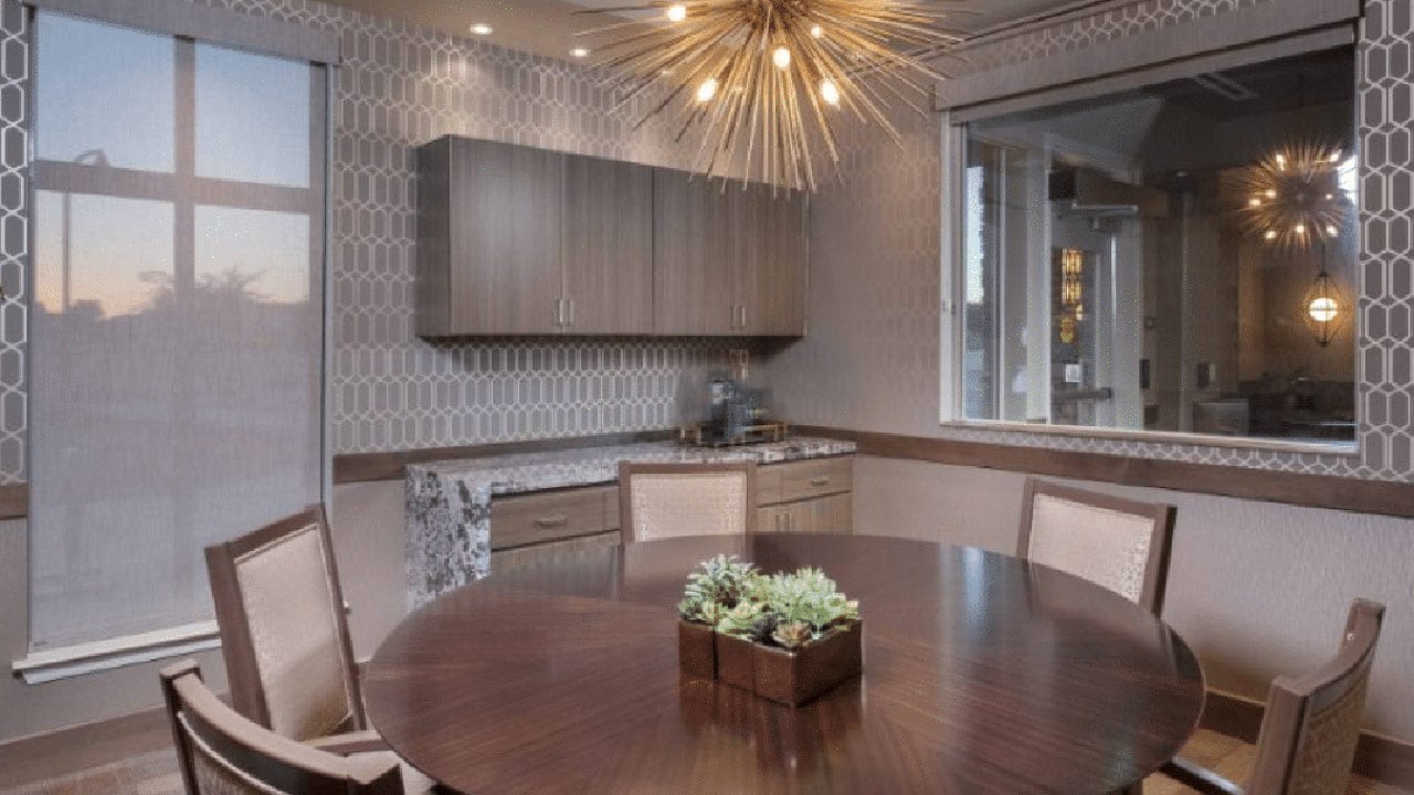 A modern dining room with a round wooden table surrounded by four chairs. A geometric light fixture hangs above. The room features patterned wallpaper, a window with blinds, and a countertop with cabinets. A small succulent centerpiece is on the table.