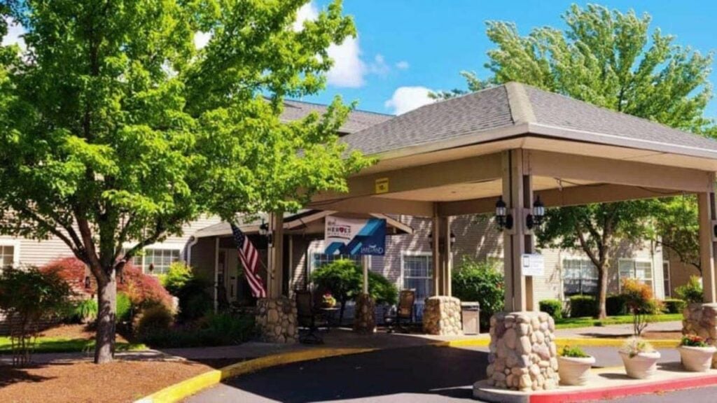 Entrance of a building with a covered driveway, surrounded by trees and landscaping. An American flag is displayed near the entrance, and banners are visible. The sky is blue with scattered clouds.