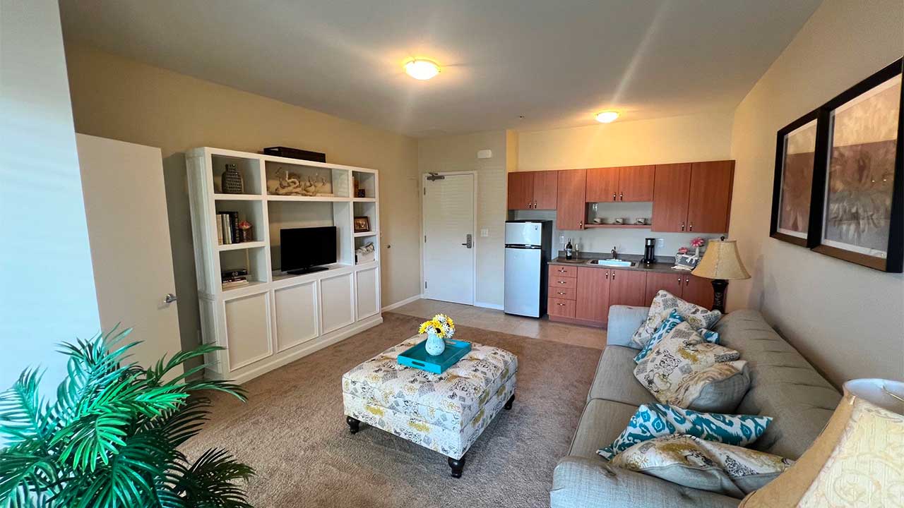 A cozy living room with a beige sofa, patterned ottoman, and white shelving unit. A small kitchen area with wooden cabinets, mini-fridge, and microwave is seen in the background. Two decorative paintings and a potted plant add charm to the space.