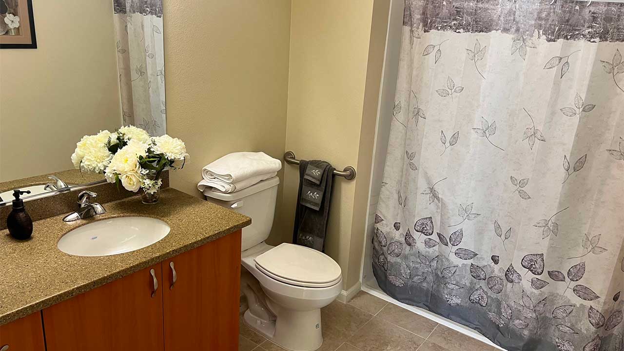 A bathroom features a sink with a granite countertop and a wooden cabinet, a toilet with folded towels on top, and a shower with a gray floral curtain. A bouquet of white flowers decorates the counter, and a black soap dispenser is nearby.