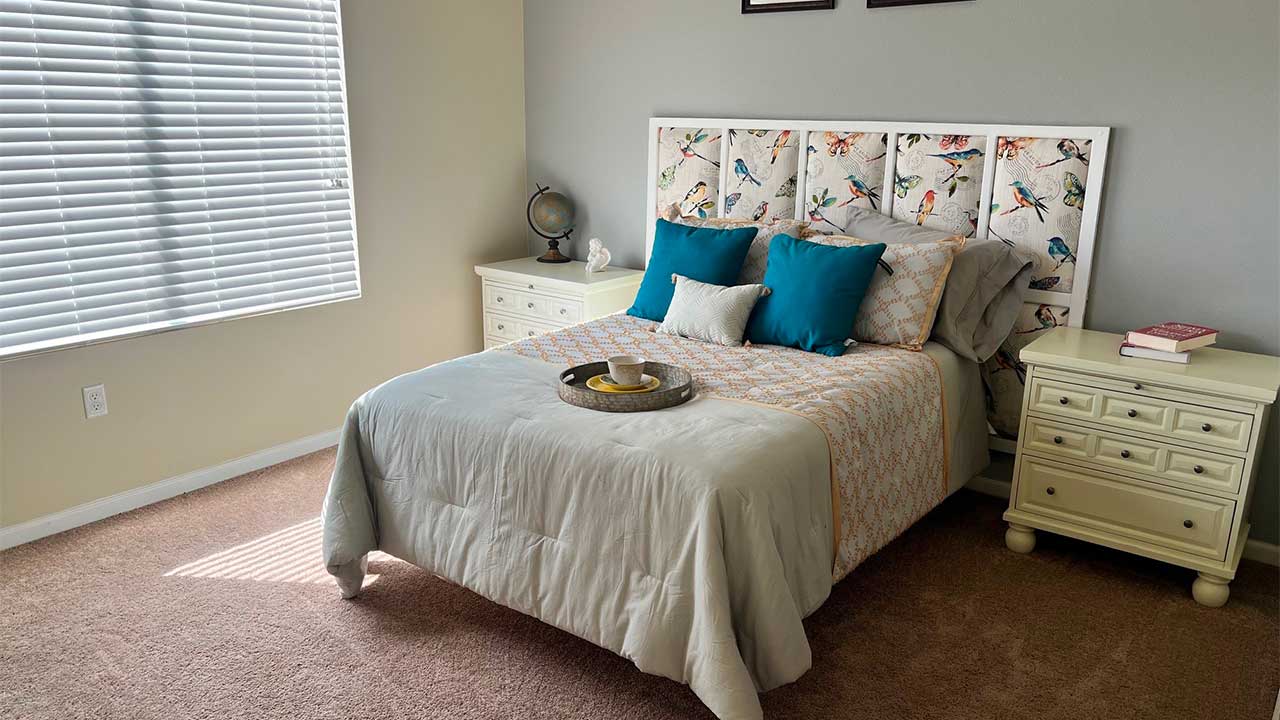 A cozy bedroom with a bed featuring a bird-patterned headboard and blue cushions. The bed is flanked by two light-colored nightstands holding books and decor. Sunlight filters through a window with blinds, casting soft shadows on the carpet.