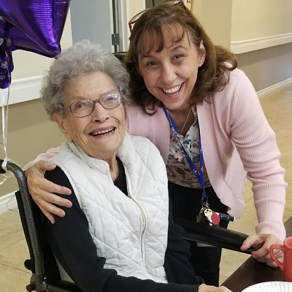 Two women smiling warmly at the camera. The elderly woman is sitting in a wheelchair, wearing glasses and a white vest. The other woman, standing beside her, has brown hair and is wearing a pink cardigan. Purple balloons are in the background.