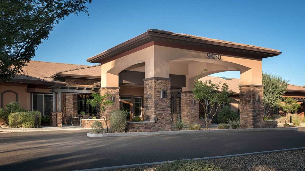 A modern building with a stone facade and arched entrance. It features large windows and is surrounded by greenery, including trees and shrubs, under a clear blue sky. The address number "9450" is displayed above the entrance.