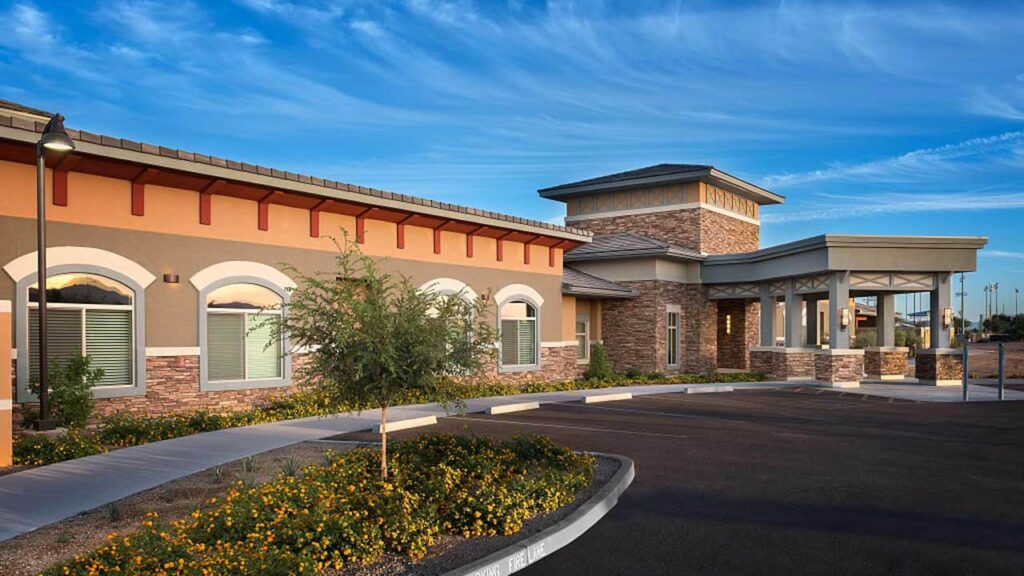 A modern, one-story building with a mix of stone and stucco exterior. It has large windows and an adjacent parking area. The foreground features trimmed shrubs and yellow flowers under a clear blue sky.