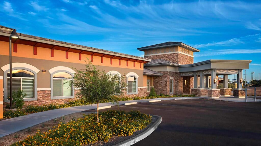 A modern single-story building with stone and brick accents sits under a blue sky. The entrance features an arched walkway. A landscaped area with yellow-flowered shrubs lines the sidewalk.