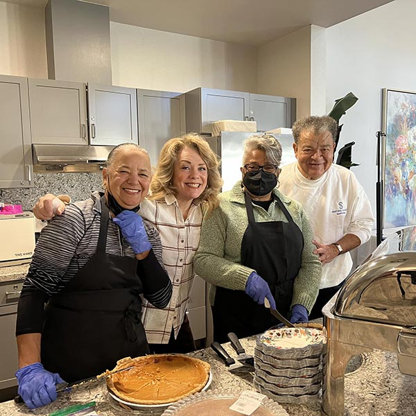Four people in a kitchen prepare desserts, with pies and serving dishes on the counter. They are smiling and wearing aprons, and one person is cutting a pie. The kitchen has modern appliances and brightly colored artwork in the background.