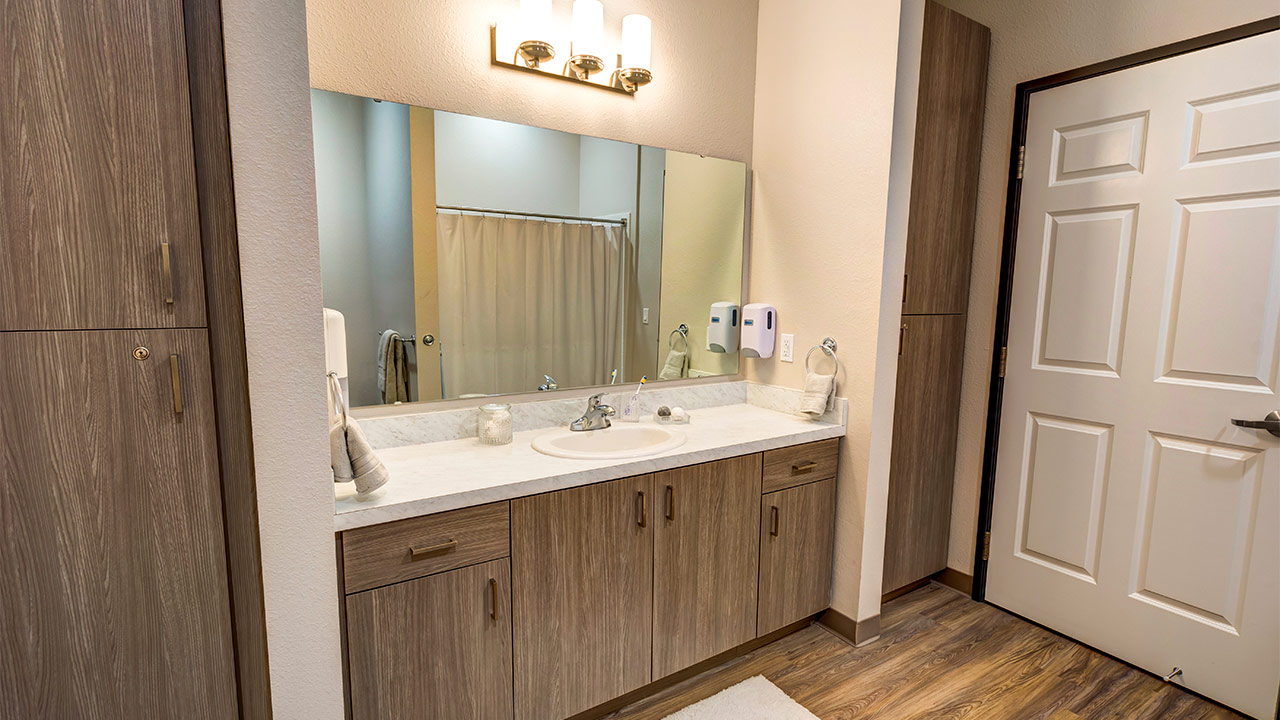 Modern bathroom with a large mirror above a wooden cabinet and white countertop. A sink is centered with toiletries nearby. The room features warm lighting, a wooden floor, closed closet, and a door leading to a shower area with a beige curtain.