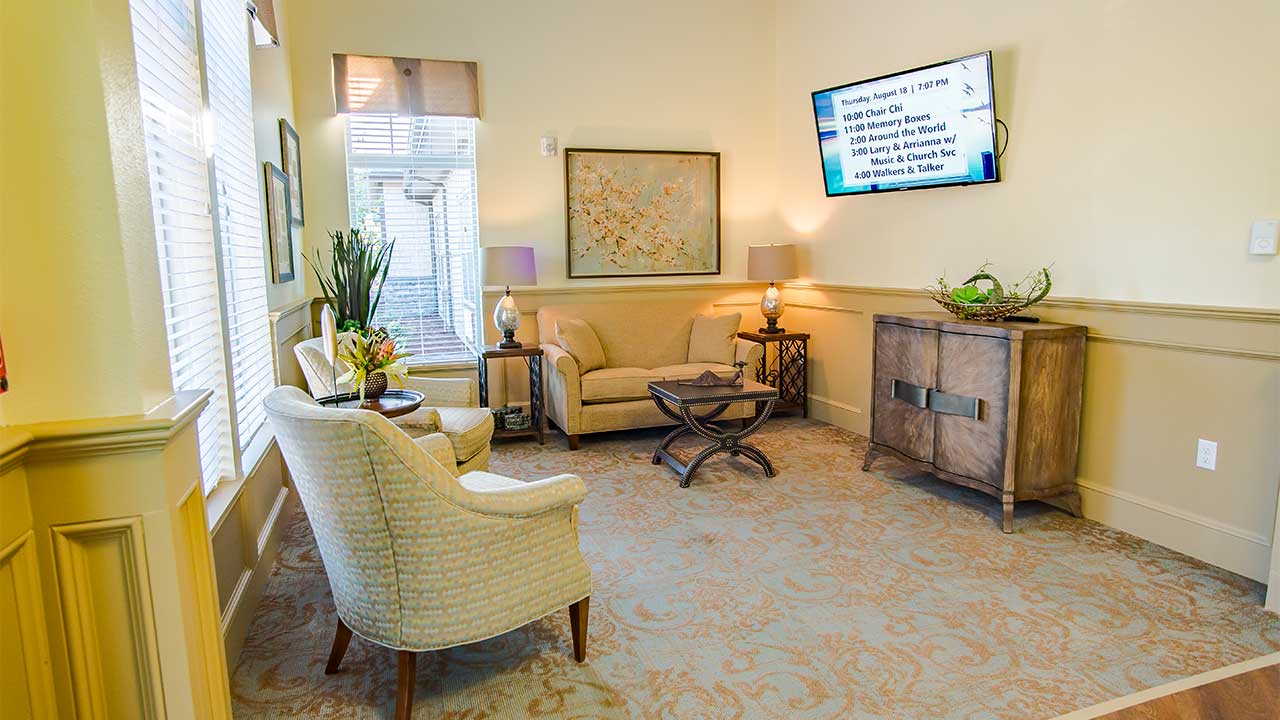 A cozy living room with two armchairs, a sofa, and a small table on a patterned rug. A painting hangs on the wall above the sofa, and a TV displaying text is mounted on the adjacent wall. Natural light filters in through a window with blinds.