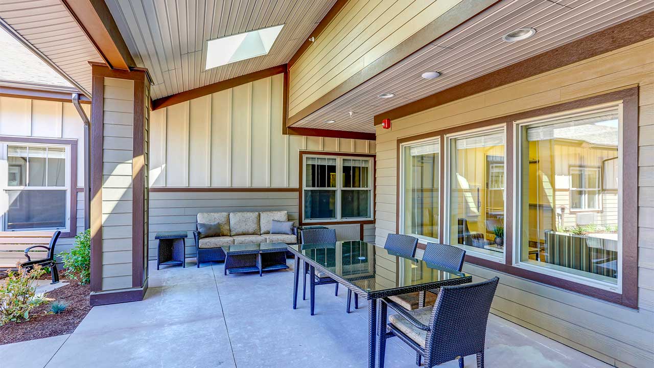 Covered patio with a skylight features a glass dining table surrounded by wicker chairs and a cushioned outdoor sofa against the wall. The area is enclosed by windows and walls of the house, with a small garden visible to the side.