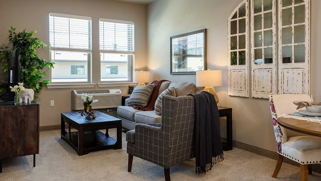 A cozy living room with a gray sofa, two cushioned armchairs, and a wooden coffee table. Lamps flank the sofa, and plants decorate the room. A window AC unit is under large windows, and wall decor includes a rustic mirror panel. A small dining table is visible.
