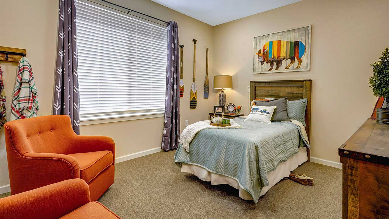 A cozy bedroom with a twin bed featuring a light blue quilt and bear-themed decor. An orange armchair sits by a window with patterned curtains. A wooden dresser and rustic items like oars and a bear painting add charm to the space.