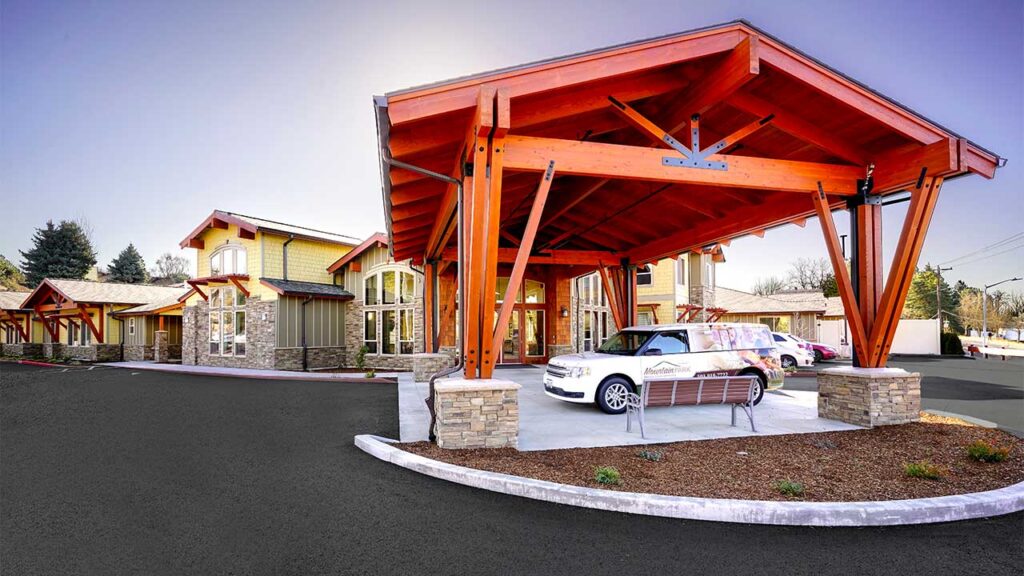 A modern building with a wooden canopy entrance and stone accents. A white car is parked under the canopy. The surrounding area is landscaped with trees and a curved driveway.