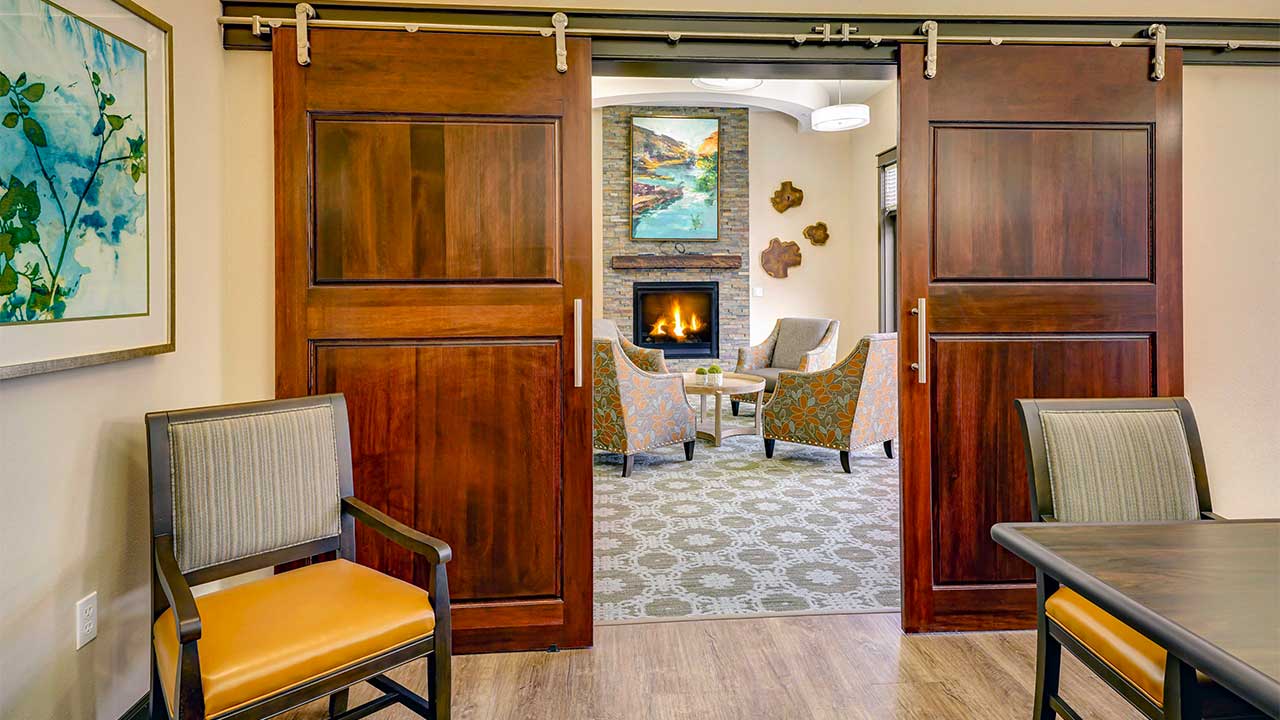 A cozy room with a modern fireplace, surrounded by four patterned chairs and a small wooden table. The room is accessed through wooden sliding doors and is decorated with paintings and warm lighting.
