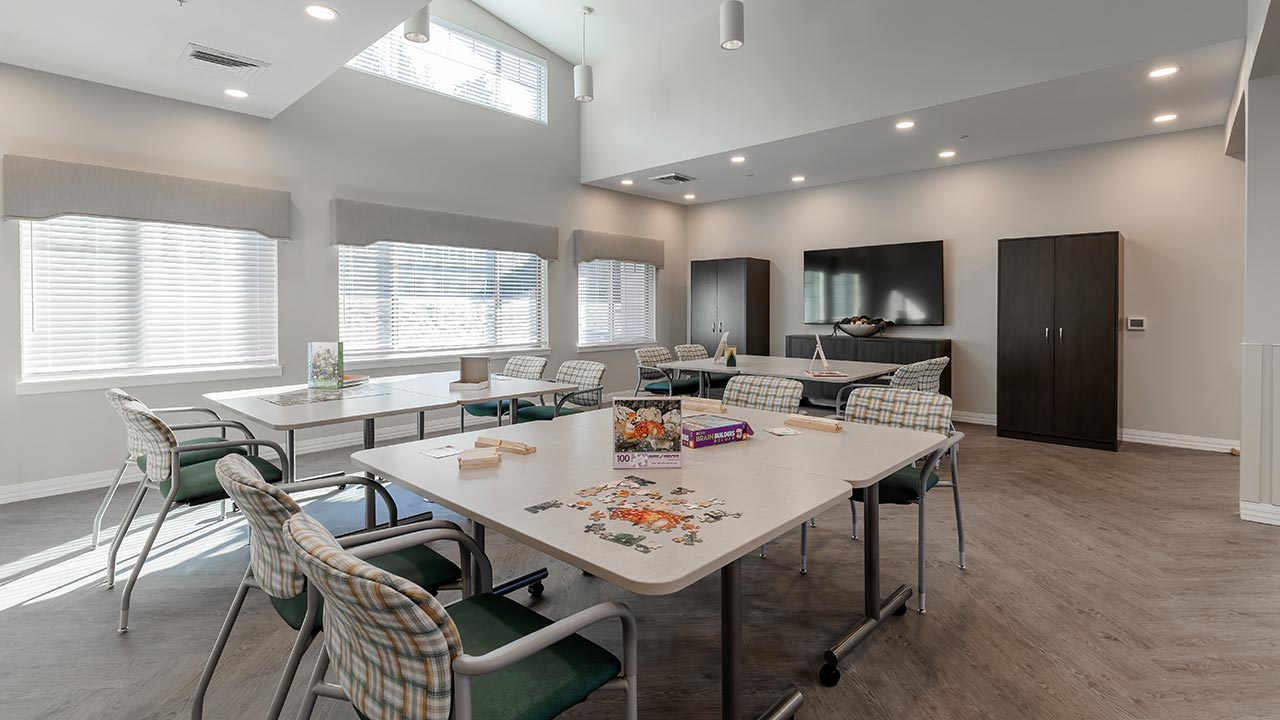 A well-lit room with large windows, several tables, and cushioned chairs. A partially completed puzzle and board games are on the tables. There's a TV and cabinets against a wall, with a light wood floor and recessed ceiling lights.