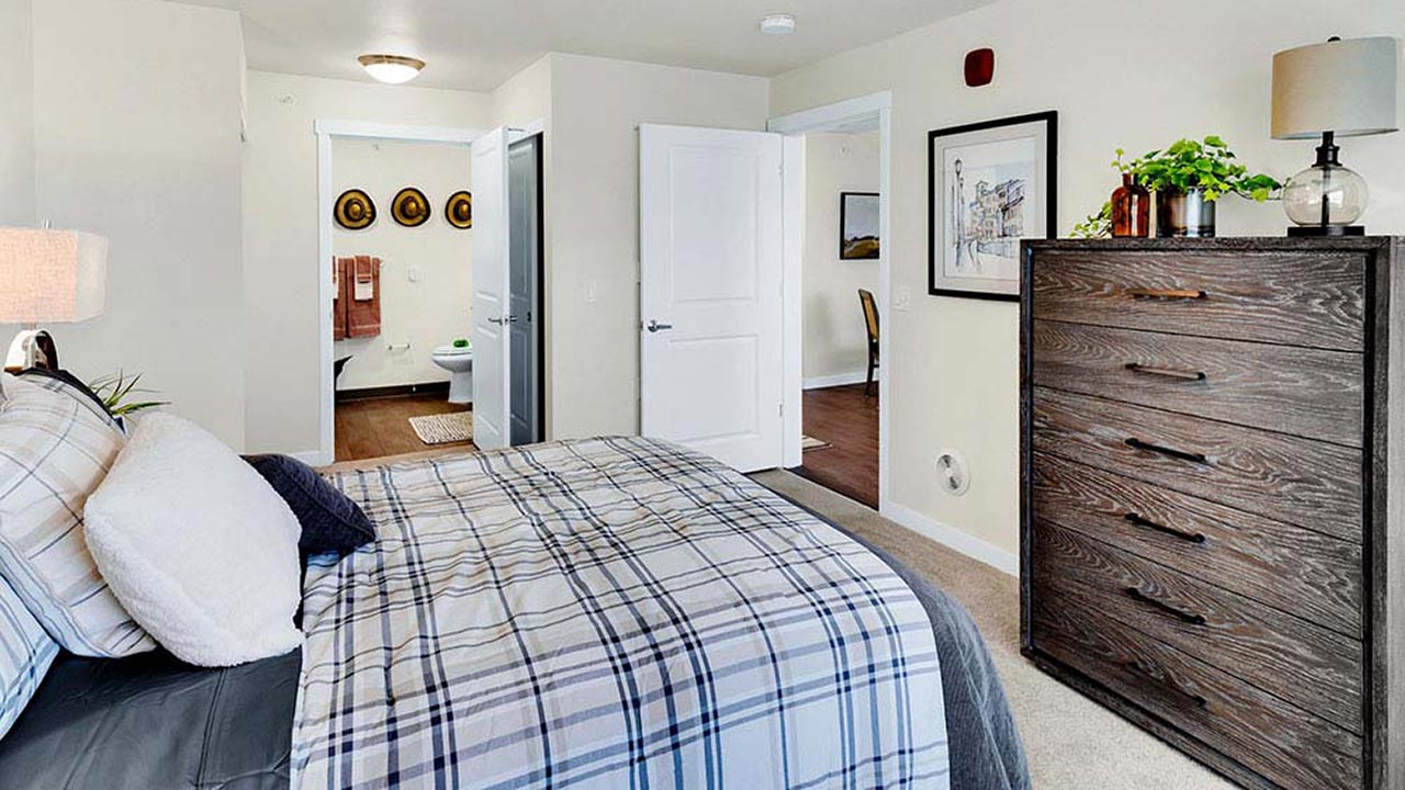 A neatly arranged bedroom features a bed with plaid bedding, a wooden dresser with plants and a lamp, and artwork on the wall. The room's doorways lead to a bathroom and another hallway. Warm lighting creates a cozy atmosphere.