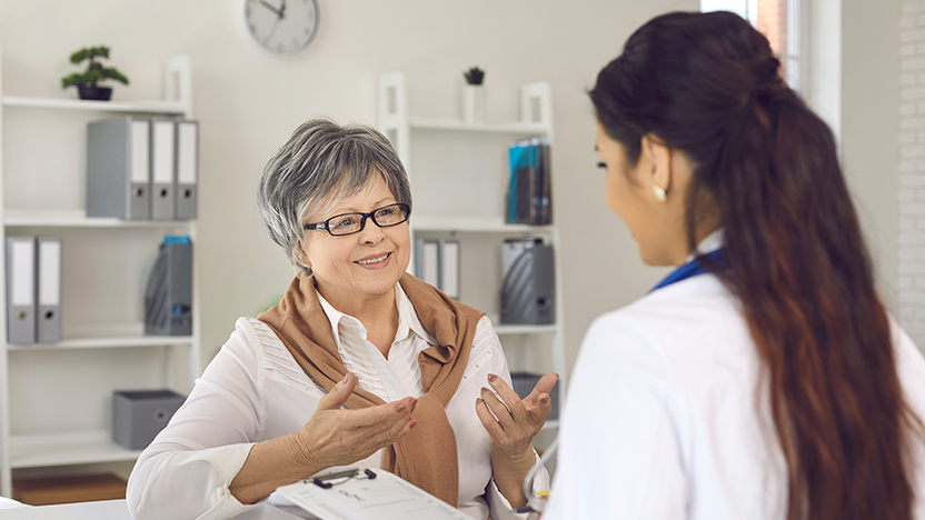 A woman with short gray hair is engaged in conversation with a female doctor in a medical office. The doctor holds a clipboard. The background includes shelves with binders and a small plant, and a clock on the wall.
