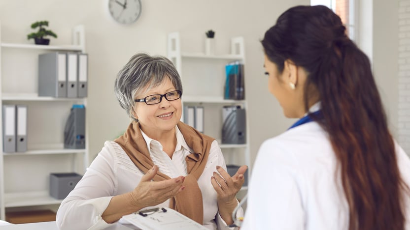 An older woman with gray hair talks to a female doctor in an office, gesturing with her hands while the doctor listens and takes notes. Shelves and a clock are visible in the background.