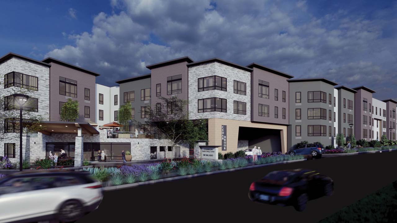A modern multi-story apartment building with white and gray stone exterior, large windows, and landscaping in front. Cars drive past on a road, and people are walking near the entrance under a cloudy sky.