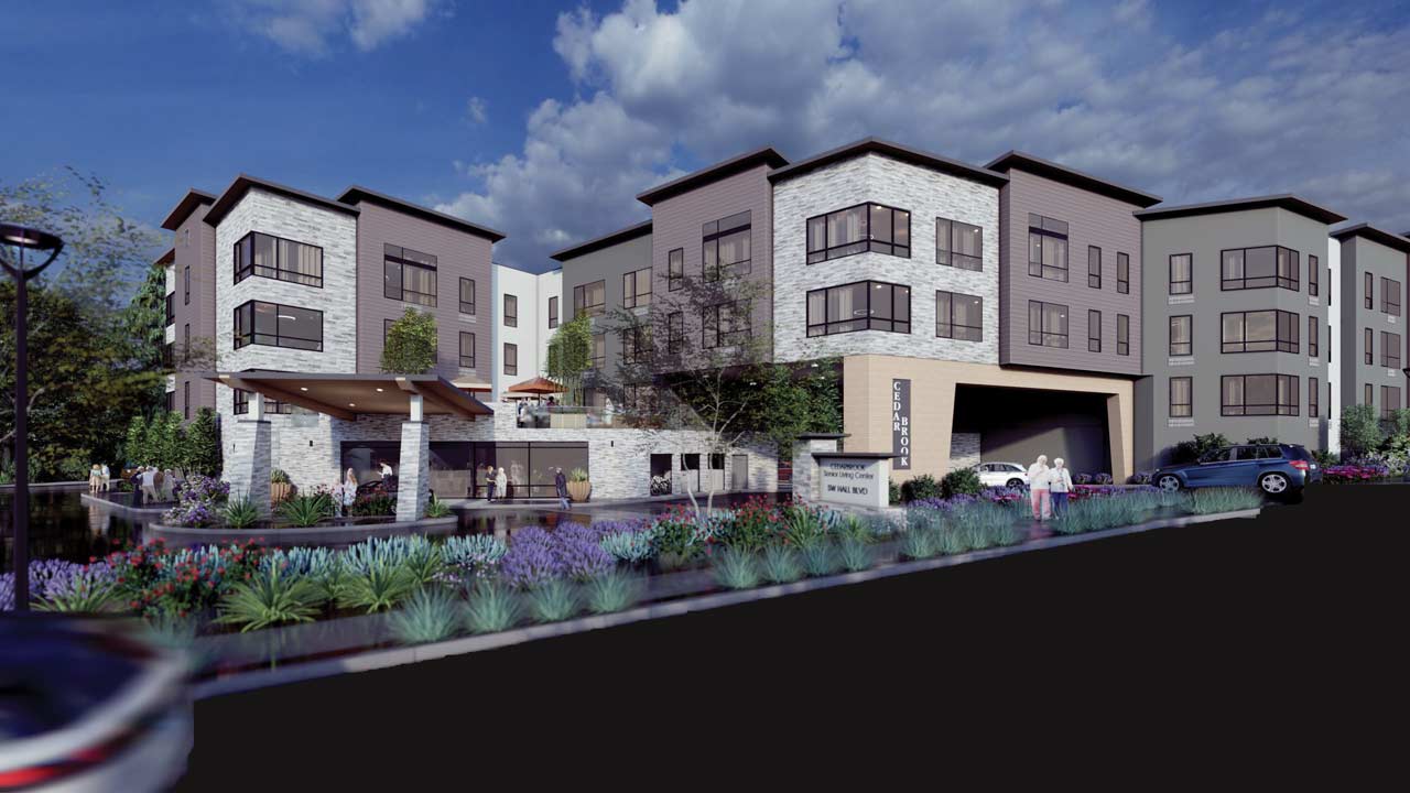 Modern apartment complex with four-story buildings, large windows, stone and wood facades, landscaped gardens with purple flowers, a swimming pool, and people walking near the entrance under a blue sky with scattered clouds.