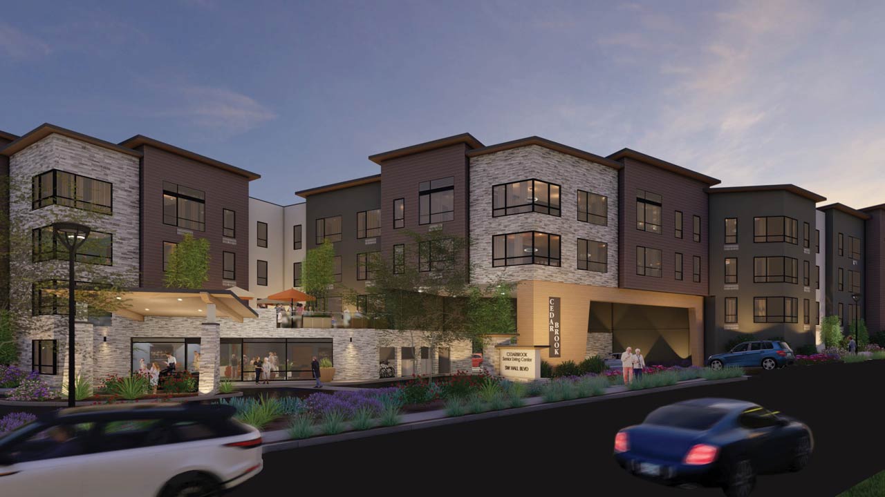 A modern three-story apartment building with large windows, a stone and wood facade, outdoor seating areas, greenery, and cars driving on the street at dusk.