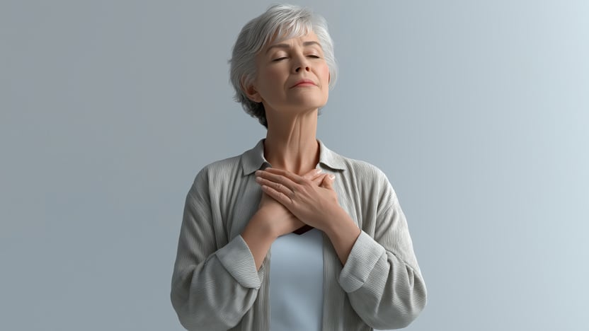 An elderly woman with short gray hair stands against a plain background, closing her eyes and resting both hands on her chest, embodying the calm and peaceful spirit often found in senior living.