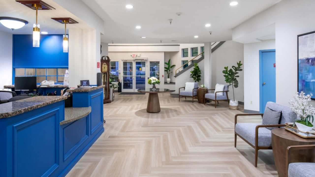 Modern lobby with blue reception desk, wood herringbone floor, seating area with gray chairs, round table with flowers, plants, staircase, and artwork on the walls. Bright lighting and large windows in the background.