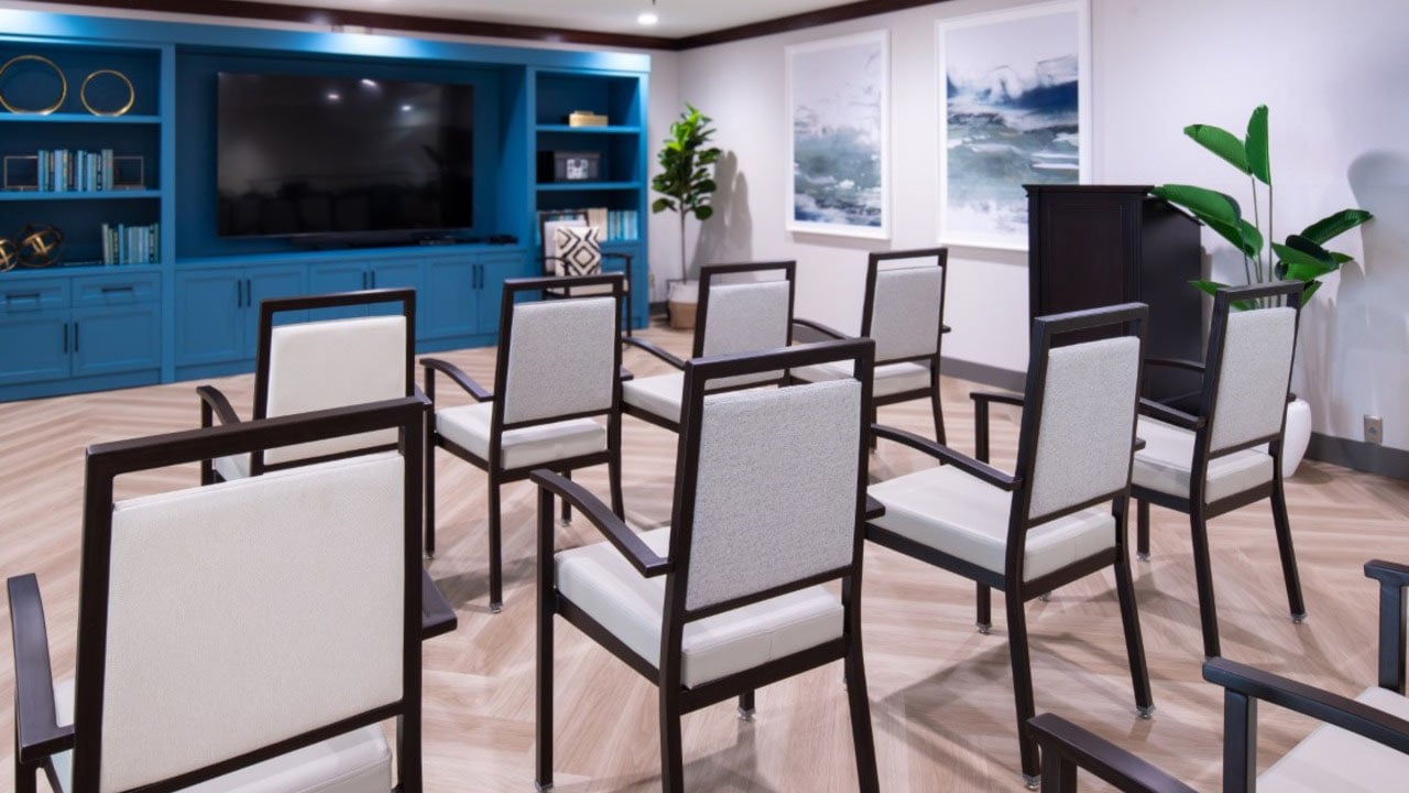 Ten empty chairs face a large TV in a modern room with blue cabinets, bookshelves, two wall art pieces, and green plants. The wood floor has a herringbone pattern.