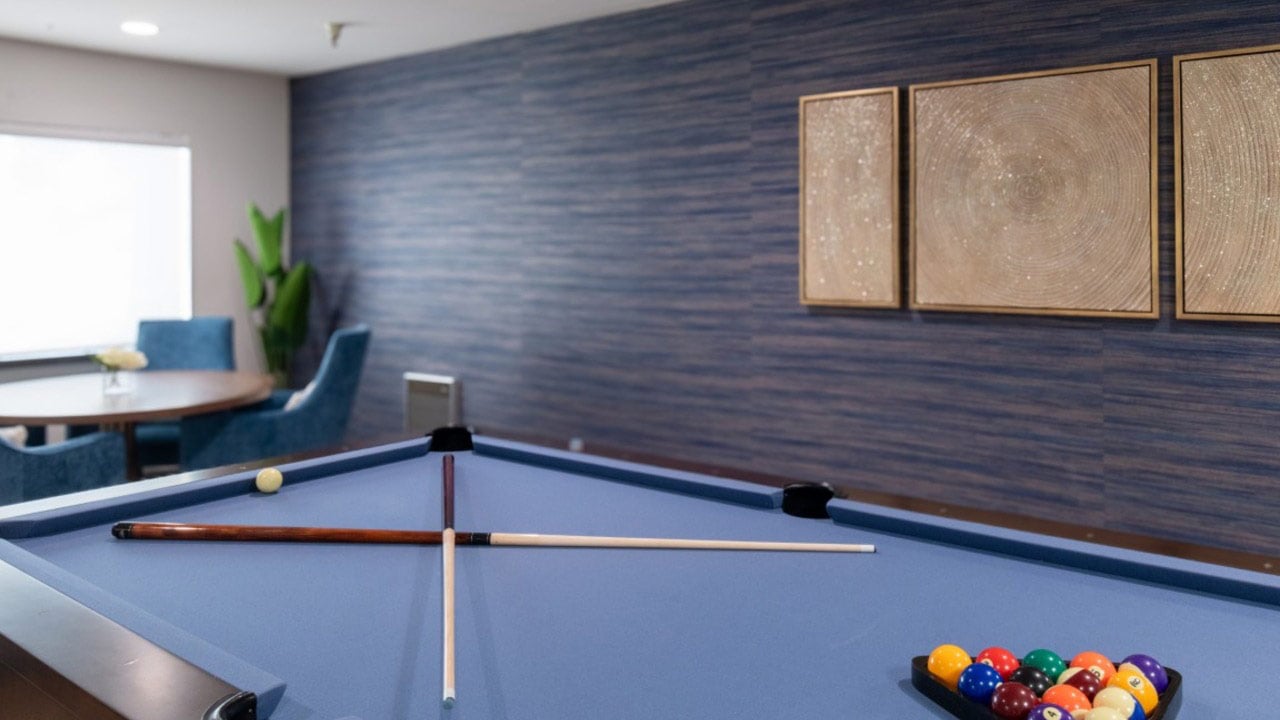 A blue pool table with cue sticks forming an X and a cue ball on top. Racked balls and triangle are in the corner. In the background, a seating area, plants, and three framed art pieces are visible on a blue textured wall.