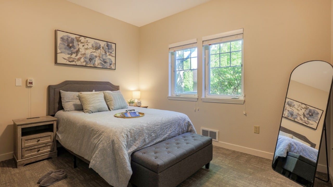 A cozy bedroom with a gray bed, blue bedding, a nightstand, a bench, wall art, a floor mirror, and two windows showing greenery outside. A pair of slippers is on the carpet near the bed.