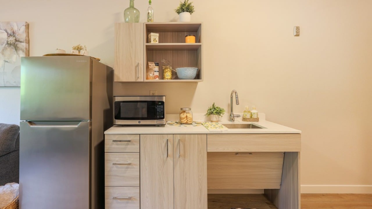 Compact kitchen with a stainless steel fridge, microwave, wooden cabinets, sink, countertop decor, and open shelves with dishes and food items against a light wall.